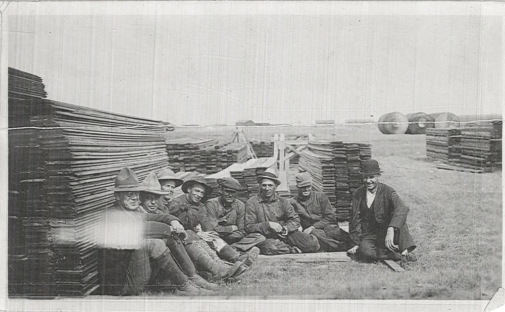 A group of men sit near stacks of lumber that would be used as concrete forms. Note the tanks in the background. 