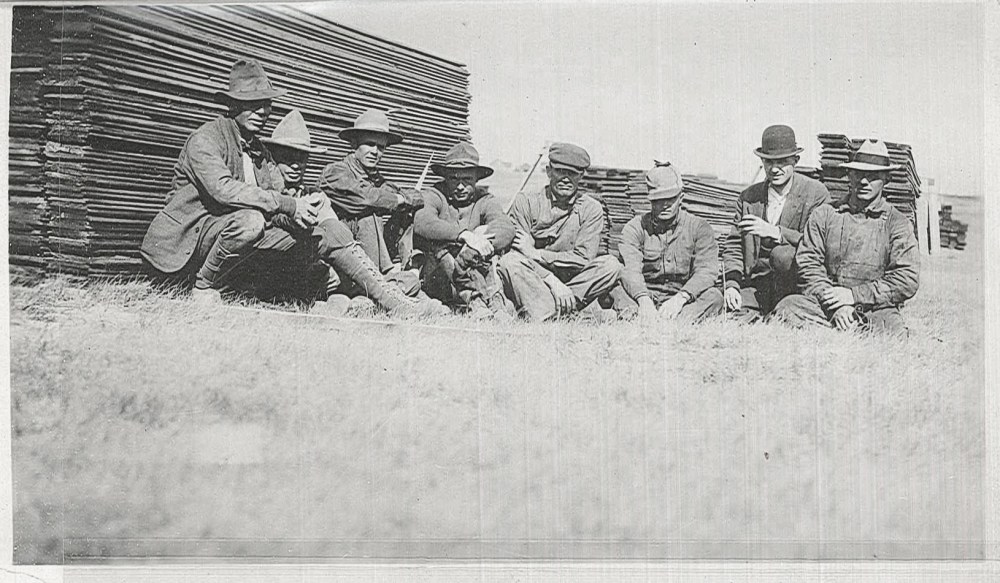 A group of men sit near stacks of lumber that would be later used as forms for concrete.  